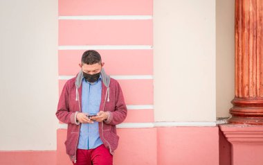 Guy with face mask checking his cell phone, young man leaning on the wall checking his cell phone