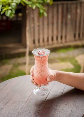 Masada çilekli milkshake 'i tutan el görüntüsü, ahşap masada çilekli meyve püresi gösteren eller. Elleri ahşap masada, arka planda bulanık çilekli smoothie tutuyor.