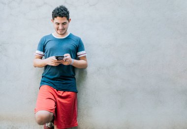 Akıllı telefon kullanarak duvara yaslanan mutlu adam. Duvara yaslanmış gülümseyen genç bir adam mesaj gönderiyor.