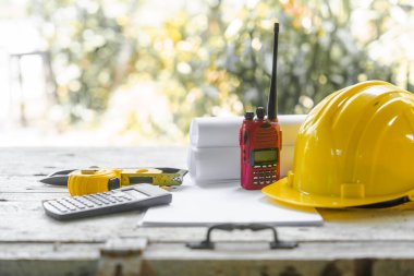 Sarı güvenlik kaskı ve ahşap bir masanın mimari planlarını katlamış. Bu da bir mimarın çalışma alanının muhtemelen bir inşaat alanında olduğunu gösteriyor..