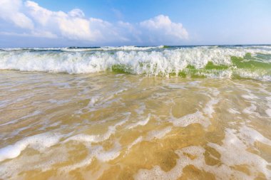 Dalgaların açık yeşil su, açık tenli kum, mavi gökyüzü ve bulutla sahile çarptığı düşük makro görüntü..