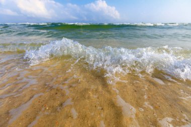 Dalgaların açık yeşil su, açık tenli kum, mavi gökyüzü ve bulutla sahile çarptığı düşük makro görüntü..