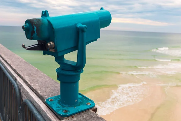 Light blue coin operated telescope mounted on the Pensacola fishing pier overlooking the beach on a partly cloudy spring day.