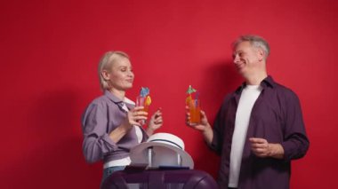 Tatil heyecanı. Mutlu bir çift. İlham perisi. Orta yaşlı kadın ve erkek dans ediyor ve kırmızı arka planda bavulun yanında yeni kokteyller içiyor..