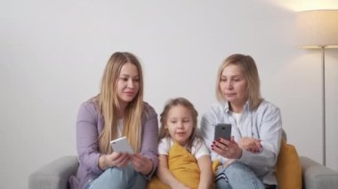İnternetten bilgi. Şaşırmış bir aile. Beklenmedik haberler. Mutlu kadın ebeveynler, iç mekan aydınlatma odasında oturan sevimli, şok olmuş kız cep telefonlarını gösteriyor..