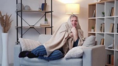 Soğuk yaşlı adam. Isıtma sistemi. Düşük ateş. Rahat, pijamalı yaşlı bir erkek kanepenin iç tarafında kareli kumaş giyiyor ve titriyor..