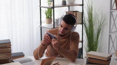 Dijital bağımlılık. Oyuncu adam. Sıkıcı eğitim. Kulaklıklı sıradan bir adam aydınlatma odasındaki kitaplarla akıllı telefon oynuyor..