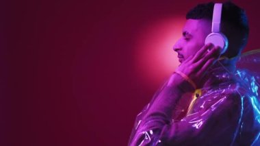 Müzik zevki. Siberpunk adam. Stereo teknolojisi. Yağmurluklu ve kulaklıklı hippi profilli adam kırmızı mor ışıklı arka planda şarkı dinliyor..