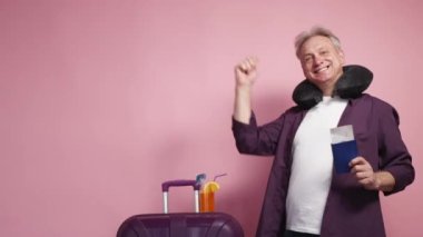 Tatil neşesi. Heyecanlı adam. Trip kutlaması. Pembe arka planda yabancı pasaport biletiyle valizin yanında dans eden mutlu orta yaşlı adam..