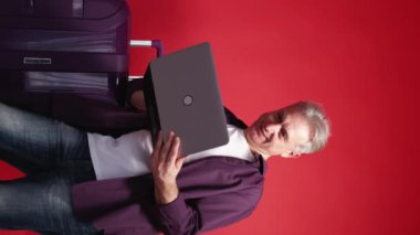 Dikey video. Çevrimiçi bağlantı. Mutlu adam. Tatil gezisi. Kırmızı arka planda bavulun yanında dizüstü bilgisayarı yazan orta yaşlı bir adam var..