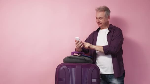 移动连接 令人惊讶的人 旅程的兴奋 快乐的中年男人摆着智能手机的姿势 提着手提箱和粉色背景的旅行用品 — 图库视频影像