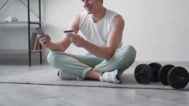 Spor dersi. Mutlu adam. Cep telefonu ödemesi. Elinde kredi kartı olan, ışık odasının iç tarafında halterlerle oturmuş akıllı telefon yazan gülen bir adam..