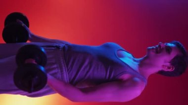 Dikey video. Kaslı adam. Neon ışıklı portre. Güçlü, mutlu adam sarı arka planda dambıllarla egzersiz yapıyor..