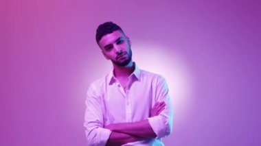 Zekice bir görünüş. Kendine güvenen bir adam. Neon ışıklı portre. Beyaz tişörtlü yakışıklı esmer adam ellerini göğsüne koyup pembe yeşil arka plan pozu veriyor..