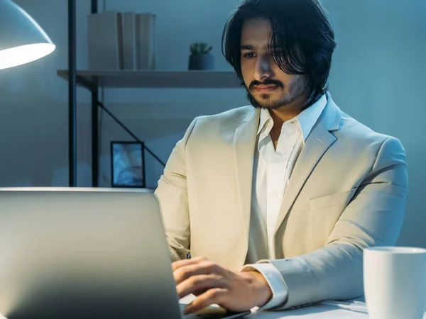 Late work. Smiling man. Project inspiration. Handsome guy in elegant look sitting desk typing laptop dark light room interior.