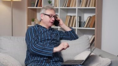 Dijital bağlantı. Modern yaşlı adam. Sanal yaşam. Pijamalı, akıllı telefonlu, dizüstü bilgisayarı daktilo eden kanepede oturan mutlu bir erkek..