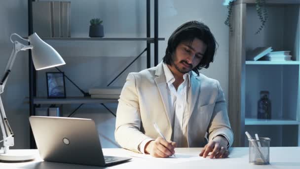 ビジネスプロジェクト 男性主任 遅い仕事だ エレガントな笑顔男でスーツの書き込みで紙文書に座ってデスクで暗い光の部屋のインテリア — ストック動画