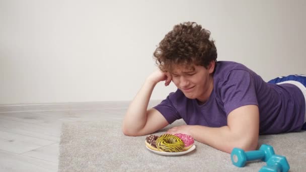 饮食问题 沮丧的男人 减肥运动 一个心烦意乱的家伙看着甜甜圈盘铺在地板上 里面有哑铃灯房间 — 图库视频影像