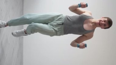 Dikey video. Spor eğlencesi. Heyecanlı adam. Saçma bir eğitim. Terli yelek giyen mutlu funky adam ışık odasının iç tarafında halterlerle zıplayan aptalı oynuyor..