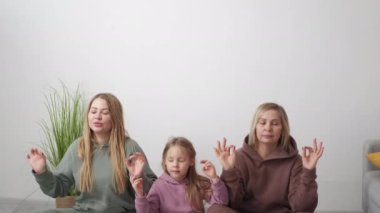 Aile meditasyonu. Kadın nesli. Sakin bir ruh hali. Yogada oturan küçük sakin kız anne ve büyükannesinin aydınlık odasının içinde gözleri kapalı poz veriyor..