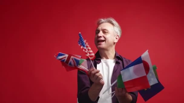 全球旅行 一路顺风假期快乐 有灵感的中年男子举着许多国家的国旗 在红色背景下跳舞 动作缓慢 — 图库视频影像