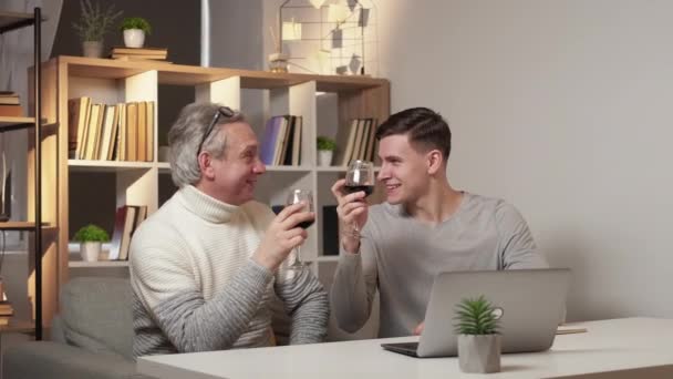 家族のお祝い 肯定的な男性 ホームレジャー 幸せな父と息子のワインを飲む肯定的なニュースを得るシッティングデスクオープンラップトップで光の部屋のインテリア — ストック動画