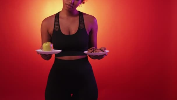 艰难的选择 多疑的女人饮食问题 瘦小的女士拿着装有苹果和不健康饼干的盘子 呈霓虹灯浅黄底色 — 图库视频影像