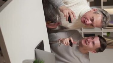 Dikey video. Online kutlama. Mutlu adamlar. Ev toplantısı. Şarap içen baba ve oğul laptopta kadeh kaldırıyor..