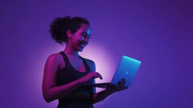 Çevrimiçi fitness. Mutlu kadın. Dijital teknoloji. Spor kıyafetleri giymiş, dizüstü bilgisayarda el ele tutuşmuş, neon ışığı, mor beyaz arka plan pozu veren pozitif bir kadın..
