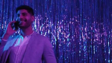 Mobil bağlantı. Mutlu adam. Neon ışıklı parti. Takım elbiseli olumlu yakışıklı adam akıllı telefondan konuşuyor. Mavi şenlikli gümüş şelale dekorasyonunda poz veriyor. Kopya alanı..