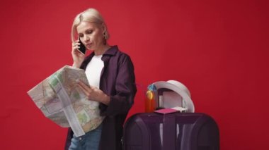 Gezici danışmanlık. Mutlu kadın. Seyahat turu. Akıllı telefondan konuşan olumlu bir bayan haritada seyahat etmek için yön seçiyor kırmızı arka planda bavulla poz veriyor.