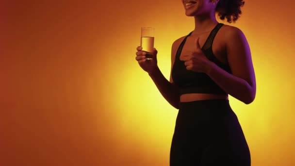 体の水分補給 協力的な女性だ 健康的なスポーツだ ネオンの光の上にポーズのようなジェスチャーを示す水の認識できない女性の保持ガラス黄色の背景 — ストック動画