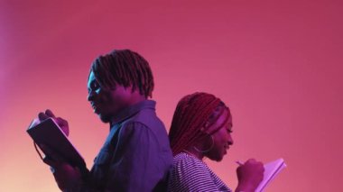 Birkaç dilek listesi. Romantik planlar. Neon ışıklı portre. Mutlu siyah adam ve kadın arkada kalıp pembe şeftali arkaplanı üzerine not defteri yazıyorlar..