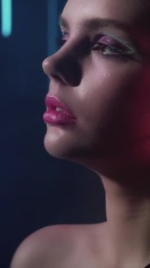Dikey video. 90 'ların modeli. Moda makyajı. Disko güzeli. Sanatsal görünümlü kendine güvenen genç bir kız yüzü. Kırmızı mavi renkte poz veriyor. Koyu renk üzerinde neon ışıklı..