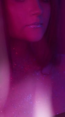 Dikey video. Neon ışıklı kız. Parlak makyaj. Gece kulübü görünüşü. Odaklanamayan mor renkli kadın yüzü, parlak tenli metalik göz farı dudaklarıyla, koyu renk üzerinde dans ediyor..