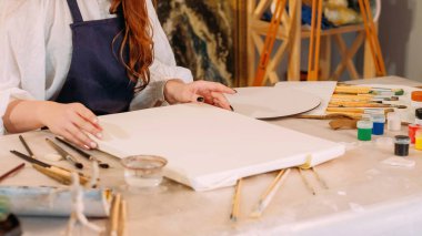 Painting hobby. Art class. Creative occupation. Unrecognizable female artist preparing blank white paper canvas at workplace with paintbrushes.