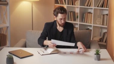 Finish work. Male analytics. Business plans. Smart concentrated man putting together documents and write in notepad sitting desk light shadow room interior.