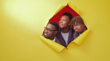 Wow effect. Curious black men. Shocked expression. Portrait of amazed male friends looking around from yellow disrupted paper on red background copy space.