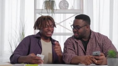 Mobile game. Excited men. Successful competition. Expressive guy boasting with high score playing smartphone showing to happy male friend light room interior.