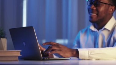 Night learning. Online research. Information technology. Cheerful smiling smart male student using laptop working late at dark night workplace.