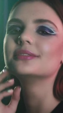 Vertical video. 90s girl. Artistic makeup. Retro fashion look. Portrait of smiling teen model face with purple color eyeshadow pink lips posing in blue neon light on dark.