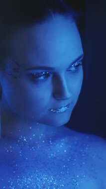 Vertical video. Sparkling makeup. Neon beauty. Disco look. Night portrait of blue color light woman face with shiny glitter skin eyeshadow lips on dark.