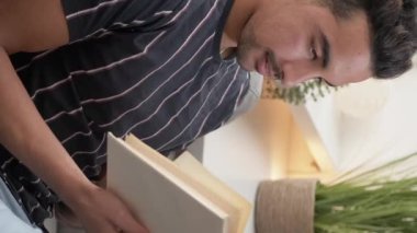 Vertical video. Enjoying reading. Curious man. Home rest. Handsome smiling guy sitting comfortable couch with interesting book in light room interior.