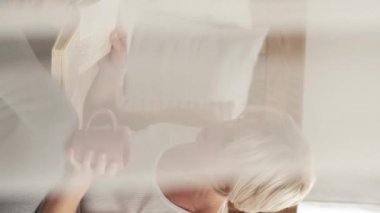 Vertical video. Home laziness. Reading woman. Enjoying life. Happy middle-aged lady drinking coffee sitting on bed with interesting book in light room interior curtain blur.