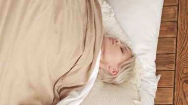 Vertical video. Leisure morning. Sleeping woman. Wonderful rest. Relaxed middle-aged lady enjoying time laying comfortable bed in light home interior.