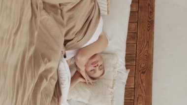 Vertical video. Enjoying morning. Awaking woman. Comfort sleep. Peaceful middle-aged lady stretching on cozy bed mattress light home interior.