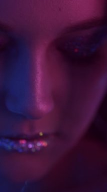 Vertical video. Neon beauty. Party makeup. Nightclub look. Closeup portrait of purple blue color light woman face with shimmering eyeshadow lips on dark.