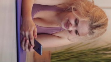 Vertical video. Phone leisure. Happy woman. Internet searching. Smiling lady in sportswear surfing smartphone laying yoga mat at home.