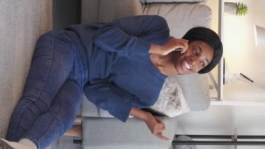 Vertical video. Positive talk. Happy woman. Home leisure. Joyful millennial lady communicating on smartphone sitting floor light room interior.