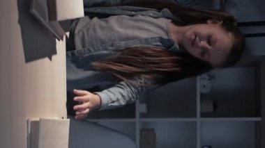 Vertical video. Kid night book. Late homework. Study overload. Tired sleepy cute little girl yawning reading at night dark home workplace interior.